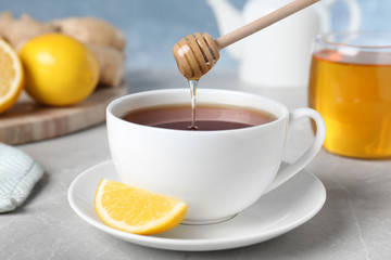 Honey pouring into cup with tea from wooden dipper on light grey marble table, closeup