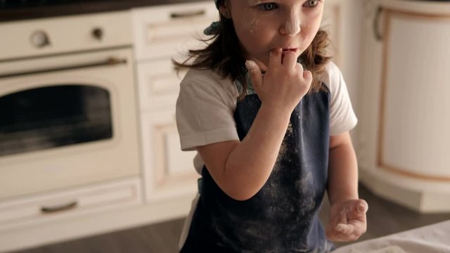 Close-up of a little girl in a cap and apron eating flour, a child helping her mother prepare cookie dough and eating raw dough and flour. The child is playing in the kitchen with food.