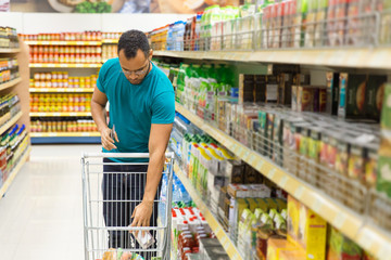 Focused African American man putting products in shopping cart. Responsible bearded guy buying food...