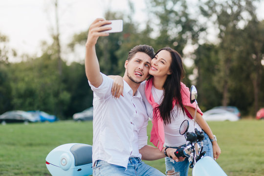 Stylish man on scooter making funny faces while taking picture with fashionable dark-haired woman. Enchanting girl with long hairstyle looking at phone screen with smile, embracing husband.