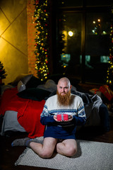Fototapeta na wymiar A man with a long red beard sits on the floor without pants in a winter sweater. man is holding a wrapped present on the background of New Year decorations and lights. Christmas tree. Parody, humor.