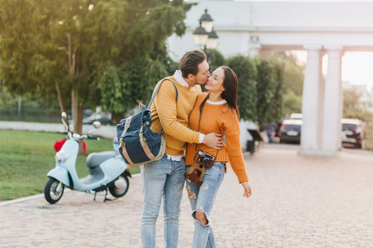 Man wears trendy jeans kissing wife during meeting in autumn park. Outdoor photo of smiling graceful girl with camera huggs with husband in weekend day.