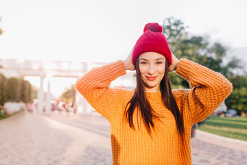 Attractive brown-haired girl in trendy orange sweater smiling on city background. Outdoor portrait...