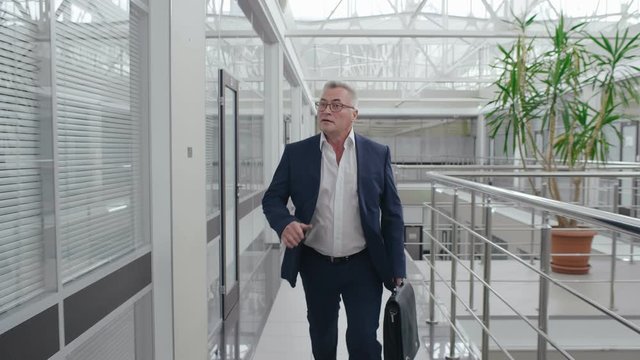 Close up shot of caucasian senior businessman in formal suit running through office hall atrium of business center in despair. Office worker is late for work and hurrying to meeting with briefcase