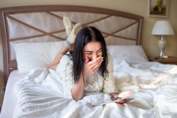 Pretty brunette girl in white knitted dress lying on bed with a tab in and hands and laughing covering her mouth with a hand