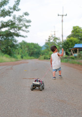 Little Asian baby boy pulls toy car walking alone on rural road. Back view.