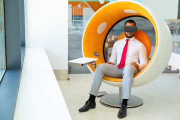 Serious young businessman using VR headset. Full length view of young African American businessman sitting in spherical chair and using virtual reality headset. Technology concept