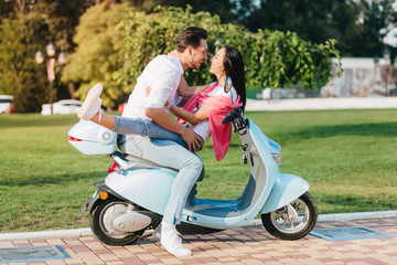 Romantic caucasian man in jeans holding girl while posing with her on scooter. Outdoor portrait of glad couple spending time together in summer weekend and smiling.