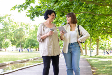 Naklejka premium Content women with tablet pc and paper cup in park. Cheerful women with coffee to go and tablet computer walking and talking together outdoors. Communication concept