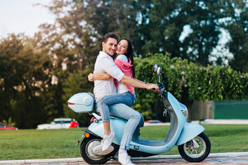 Fototapeta na wymiar Outdoor portrait of interested man having fun in weekend with pretty girl in jeans sitting on his knees. Laughing young lady posing on scooter, embracing husband during photoshoot.