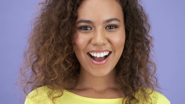 Close up view of beautiful happy african young woman in yellow t-shirt having a secret and showing silence gesture while looking at the camera purple background isolated 