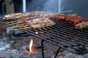 Closeup barbeque grill roasting on briquettes and charcoals, skewers, steaks and sausages in an outdoor barbecue party with flame and smoke. Blurred background, soft focus at the foreground