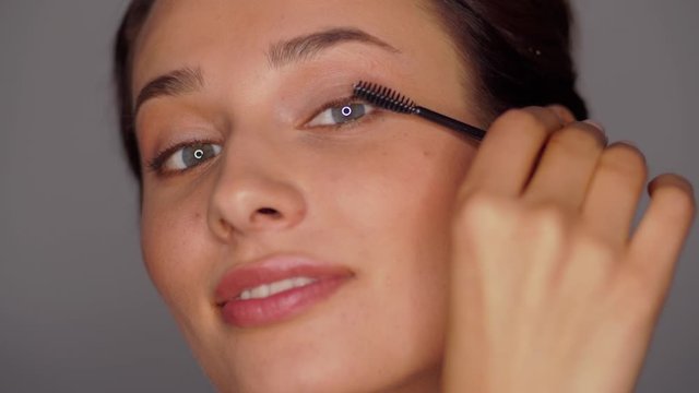 beauty, makeup and cosmetics concept - face of beautiful young woman applying mascara over grey background