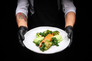 male cook in a black apron holds a plate with cooked bird fillet