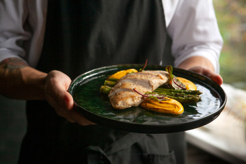 male cook in a black apron holds a plate with cooked bird fillet