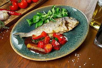 Fried fish dorado with spices and olive oil garnished with spinach, tomatoes and cucumber in sweet and sour sauce on a blue plate on a wooden background. Restaurant serving. Close up