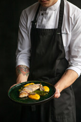 male cook in a black apron holds a plate with cooked bird fillet