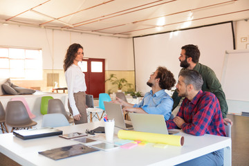 Creative male workgroup sitting in meeting room and greeting walking up female coworker. Business colleagues in casual sitting together in contemporary office space. Team meeting concept