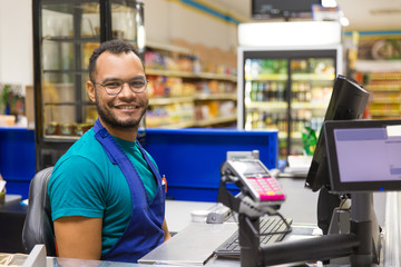 Smiling African American cashier sitting at checkout. Cheerful bearded young man in eyeglasses at...