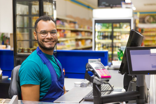 Smiling African American cashier sitting at checkout. Cheerful bearded young man in eyeglasses at workplace. Shopping concept