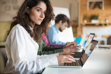 Pensive female manager using laptop at meeting table with coworkers. Business colleagues in casual working together in contemporary office space. Meeting or workplace concept