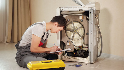 skilled repairman writes on clipboard checking broken washing machine top loader on floor in room...