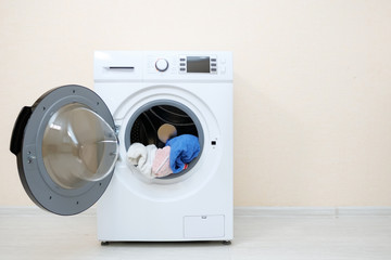 modern washing machine with heap of laundry in drum and open door stands on wooden floor near beige...