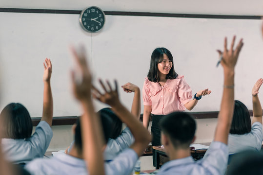 An smiling Asian female high school teacher teaches the white uniform students in the classroom by asking questions and then the students raise their hands for answers.