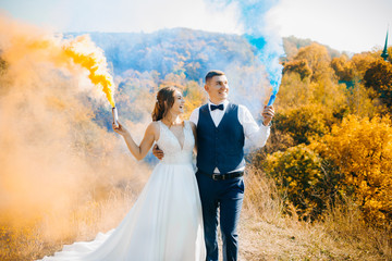 Smiling couple stands on the background of autumn forest in the color smoke. Blue and orange smoke checkers