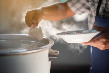 The mass of steam reflected in the morning light coming out of a large electric rice cooker that is heated in the cafeteria, where the cook is scooping the rice onto a dish.
