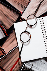 Many multicolored thick open books stand on a dark background. On the books are old round glasses and an open notebook
