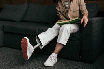 Cropped view of young woman with prosthetic leg reading book on sofa