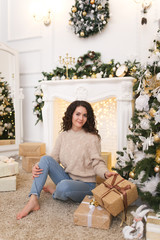 A pretty beautiful cute curly haired brunette girl in a beige sweater and jeans sits on the floor against the background of a Christmas tree and a fireplace in a spacious room decorated with garlands.