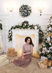 Attractive elegant beautiful brunette girl in a tight sweater and pleated skirt with a gift in her hands on the background of the fireplace, Christmas tree, garlands and bokeh in the white living room