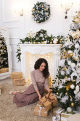 Attractive elegant beautiful brunette girl in a tight sweater and pleated skirt with a gift in her hands on the background of the fireplace, Christmas tree, garlands and bokeh in the white living room