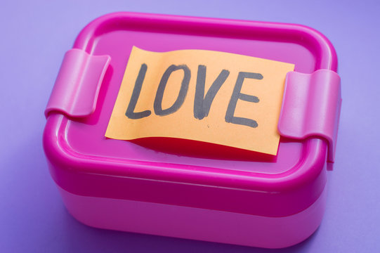 Pink Lunch Box With Orange Sticker On Purple Background. A Love Message On Sticker. Lunch At School From A Loving Wife Or Mother With Note. Bright Care Concept. Idea For Food From Home Packaging.