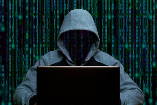 Hacker in the hood sits at a laptop in a dark room on a background of binary code