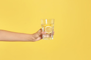 Young woman holding glass of lemon water on yellow background, closeup