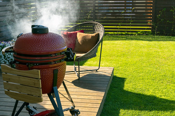 Barbecue grill and armchair in modern homes terrace. Smoke coming out of red ceramic Barbeque.