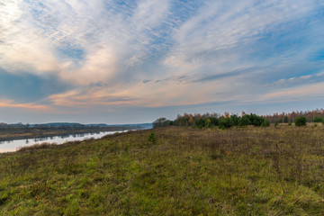 The river reflects trees and the evening sunset sky with colorful stripes of clouds above a field with wilting grass. In the distance is an autumn forest. Beautiful atmospheric landscape made at dusk