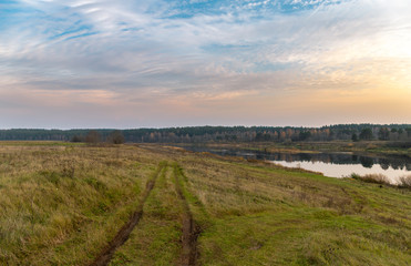 Fototapeta na wymiar Calm panoramic landscape. Along the river, a country road with a dirty traces goes into the distance. The opposite shore with trees is reflected in the water. Violet-orange clouds in the sunset sky