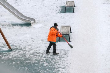 Worker in an orange jacket sweeps snow on a playground in a park