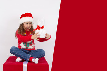 An emotional portrait of a little girl in a Santa Claus hat, sitting on a cube with a gift in her hands, on a red and white background. The concept of a happy childhood. Blank space for text. mock up