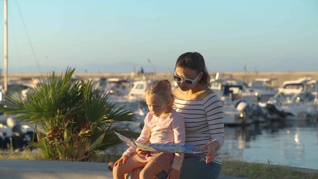 Young Woman with her Daughter Reading Tourist Map while Sitting near the Marina in Forio Town on Ischia Island, Italy. Lots of Motor Boats and Yachts Moored Nearby.