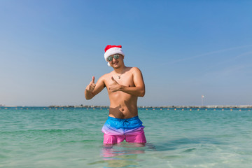 Young man in santa hat and sunglasses show thumbs up while standing at sea background