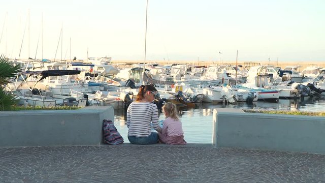 Young Woman with her Cute Little Girl Reading a Touristic Map while Sitting on the Steps near Yachts Parking in Harbor at Sunset. They Travelling around Ischia Island in Italy.