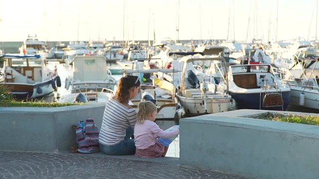 Cute Little Girl and her Mother Sitting on the Steps of the Port of Forio Town on Ischia Island. They Planning Their Trip on Island by Reading Tourist Map and Using Online Maps on Mobile Phone.