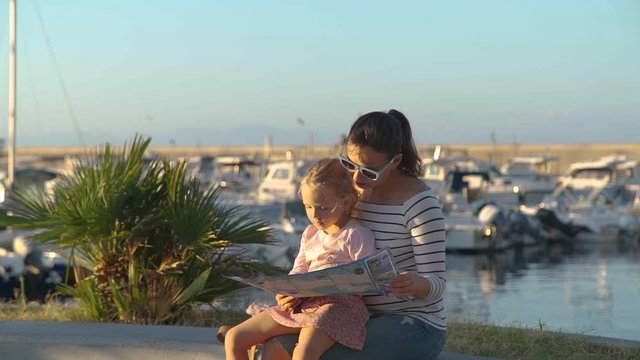 Young Woman with her Daughter Reading Tourist Map while Sitting near the Marina in Forio Town on Ischia Island, Italy. Lots of Motor Boats and Yachts Moored Nearby.