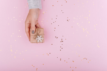 A small Christmas gift with a snowflake in Kraft paper in a woman's hand on a pink background with gold sequins