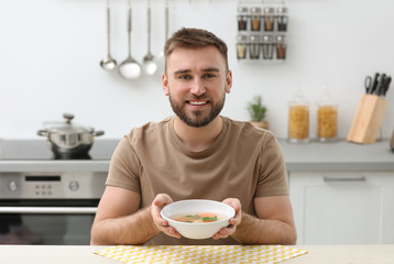 Young man holding bowl of tasty soup at table in kitchen